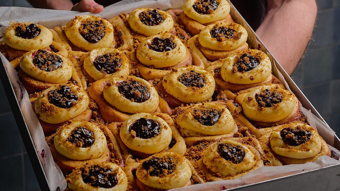 A tray of scrolls topped with cream cheese frosting, blueberry compote and biscuit crumb