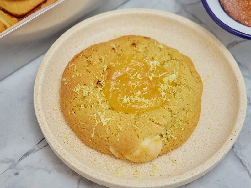 Lemon White Choc shortbread cookie on a plate beside a tray of maple and walnut scrolls